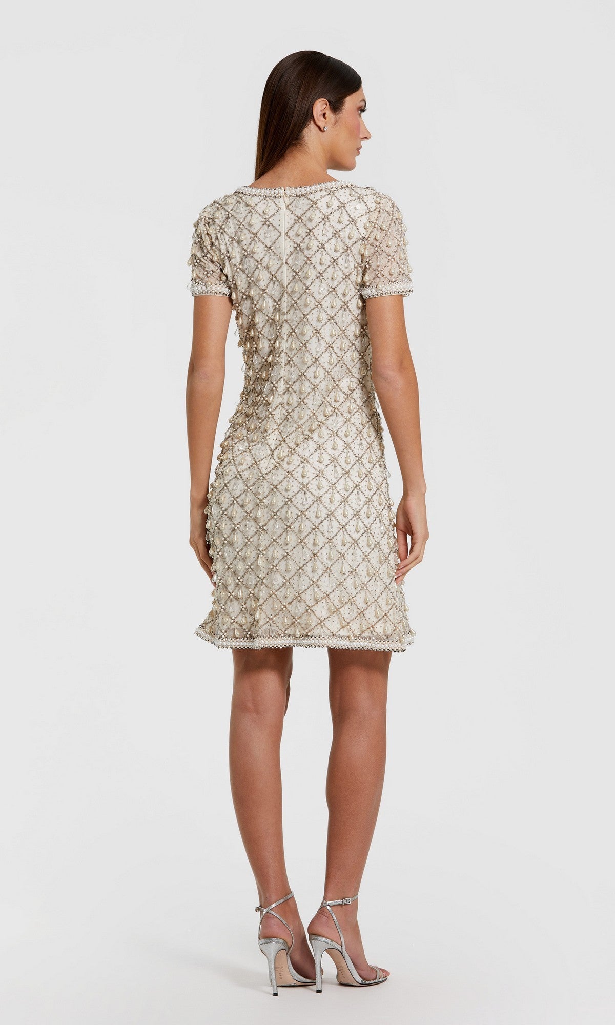 A woman faces away in a Mac Duggal 10097 short semi-formal dress featuring short sleeves, knee-length cut, and a textured diamond pattern, paired with silver high-heeled sandals against a plain background.